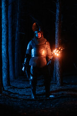 Obraz premium A knight in full armor, sword and torch in hand, walks through a gloomy night forest. A medieval warrior, bloodied and scarred, hunts and searches in the darkness.