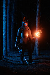 Obraz premium A knight in full armor, sword and torch in hand, walks through a gloomy night forest. A medieval warrior, bloodied and scarred, hunts and searches in the darkness.