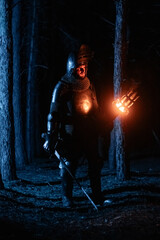 Obraz premium A knight in full armor, sword and torch in hand, walks through a gloomy night forest. A medieval warrior, bloodied and scarred, hunts and searches in the darkness.