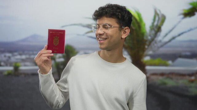 Young man smiling and holding red denmark passport on street with blurred hills in daylight; travel excitement.