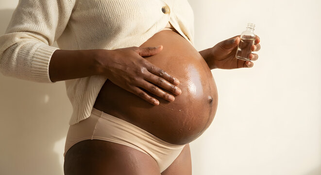 pregnant black woman applying natural body oil to her belly for stretch mark prevention and skin care during pregnancy wellness and maternity self care concept on neutral background