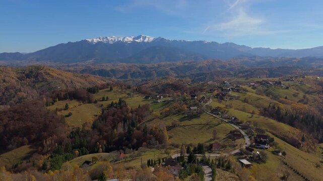 Picturesque Autumn Hillcrest Village and Driving Cars in Pestera under Snow Capped Bucegi Mountains Aerial Descending Shot 4K Graded
