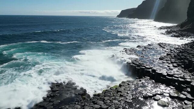 Waves crashing against hexagonal rock formations along the coastline under a bright blue sky on a sunny day
