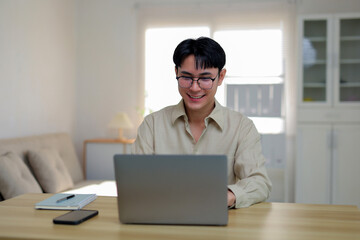 Fototapeta premium Young man wearing glasses smiling while working on a laptop computer at a wooden desk in a modern home office