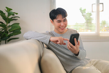 Obraz premium Young man smiling and scrolling on smartphone, connecting with people in a comfortable home environment