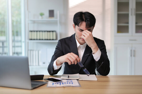 Young businessman experiencing headache and eye strain at office desk, taking off glasses due to overwork and stress