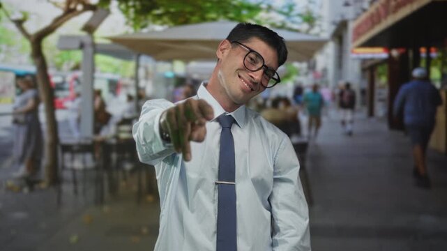 Man wearing glasses and tie points finger at camera beside sunlit cafe table on street; joyful invitation.