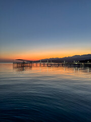 Fototapeta premium Evening sunset at Issyk Kul Lake in Kyrgyzstan. Long illuminated pier stretching into calm water with mountains on horizon.