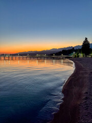Fototapeta premium Evening sunset at Issyk Kul Lake in Kyrgyzstan. Long illuminated pier stretching into calm water with mountains on horizon.