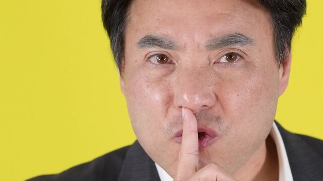 Close-up Portrait of Asian Businessman Making Silence Gesture on Yellow Background