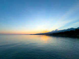 Obraz premium Sunset at Issyk Kul Lake in Kyrgyzstan. Sun rays breaking through clouds reflecting in calm water with mountains in background.
