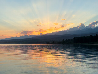 Obraz premium Sunset at Issyk Kul Lake in Kyrgyzstan. Sun rays breaking through clouds reflecting in calm water with mountains in background.