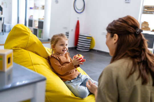 Young girl on a yellow beanbag holding an apple, smiling and talking with her child psychologist in a bright, warm playroom focused on emotional well-being and therapy