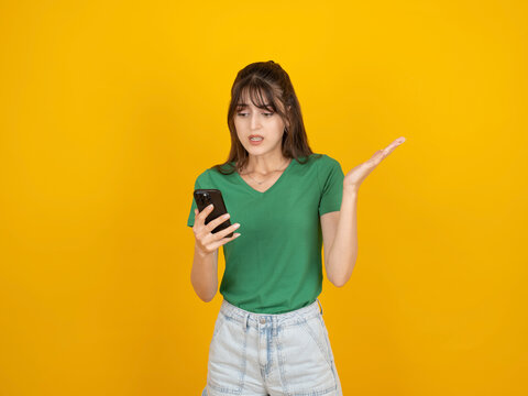 Confused young woman looking at her smartphone with a questioning gesture, standing against a vibrant yellow background. Concept of technological problems, mobile app errors, and feeling lost with dig