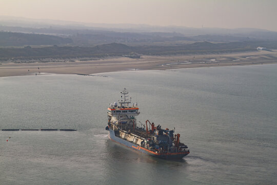 Monster, Netherlands - 13 February 2026: Aerial view of a dredger ship sailing on the grey ocean waters near the sandy coastline, fading into the misty horizon.