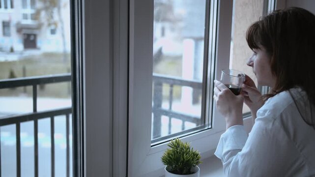 Cozy Coffee Time: Woman Enjoying a Window View and a Warm Beverage on a Gloomy Day