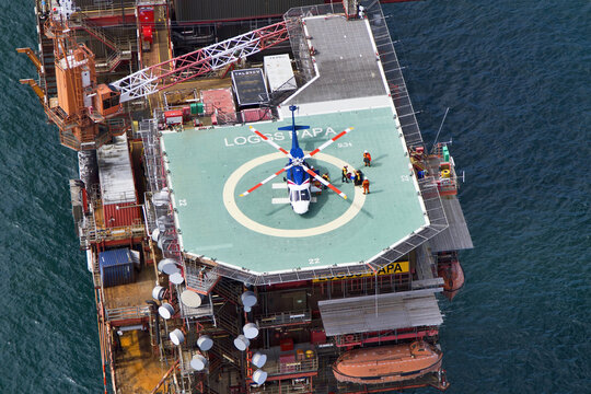 United Kingdom, United Kingdom - 15 February 2026: Aerial view of a blue and white helicopter landed on a helipad, surrounded by figures, atop a vast, complex offshore oil rig structure.