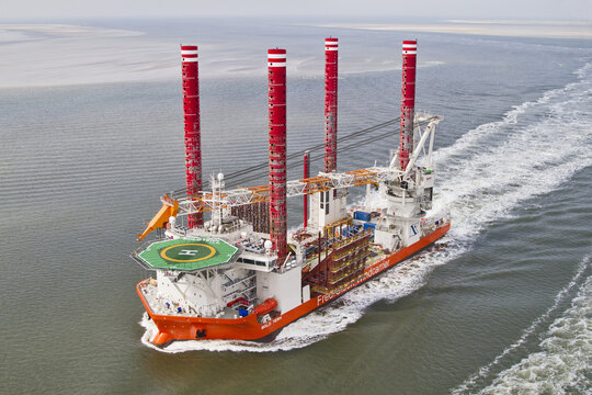 Esbjerg, Denmark - 15 February 2026: Aerial view of a massive orange and white offshore installation vessel cutting through the grey sea, its red and white striped legs reaching towards the sky.