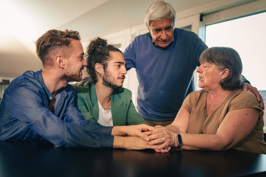 Multi-generational family sharing emotional support and empathy at home. LGBTQ+ couple with parents holding hands in a moment of resilience and connection.