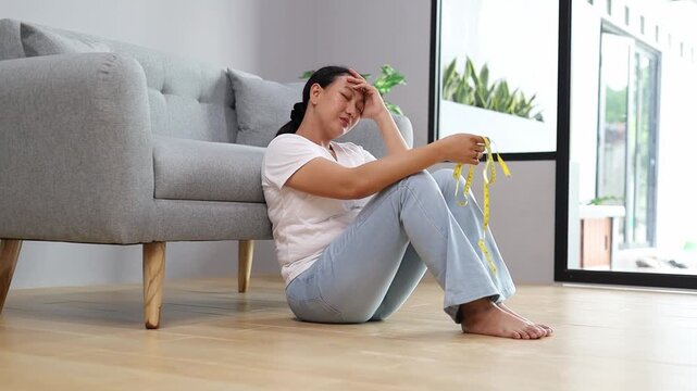 Upset Asian housewife sitting on floor at home feeling distressed with measuring tape showing body insecurity
