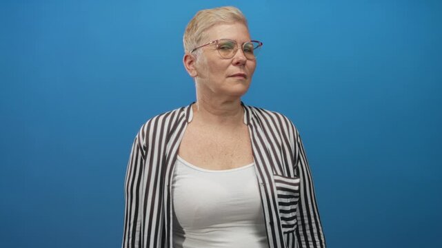 Woman with glasses turning head to side showing bare upper chest and striped shirt in blue studio; quiet confidence.