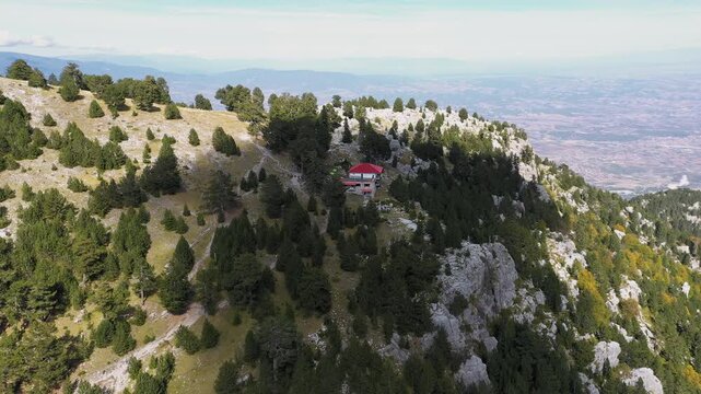 Slow panning drone shot of Spilios Agapitos Refuge nestled among fir trees in the rocky landscape of Mount Olympus under a blue sky with clouds