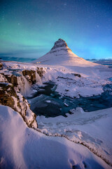 Kirkjufell Mountain Under Aurora: Majestic Kirkjufell mountain, its peak kissed by the ethereal glow of the aurora borealis, creating a symphony of color over a pristine, snow-covered landscape.