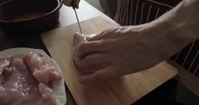 Professional chef in striped apron slicing fresh raw chicken breast on wooden board in kitchen, close up of man hands cutting poultry meat for cooking dinner at home, editorial lifestyle.