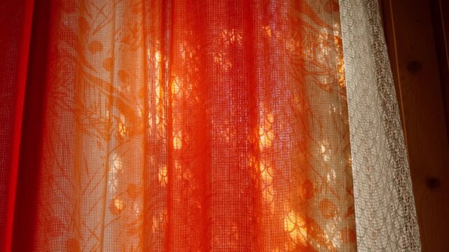 Golden hour sunlight shines through orange fabric curtains creating warm glow in rural house. Intricate lace border visible on side. Peaceful summer day interior in Cesis, Latvia.