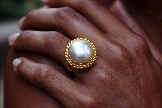 A hand shows a large pearl ring with a golden band. The background is soft and not clear