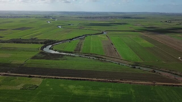 Captivating drone shot showcasing the natural curves of a waterway cutting through Moroccan farmlands. Features the expansive green landscape and the distant skyline of Larache city.