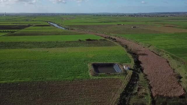 Drone shot capturing a functional farm water storage unit next to a natural reed canal. Highlights the efficient irrigation methods used in the lush agricultural plains of Northern Morocco.