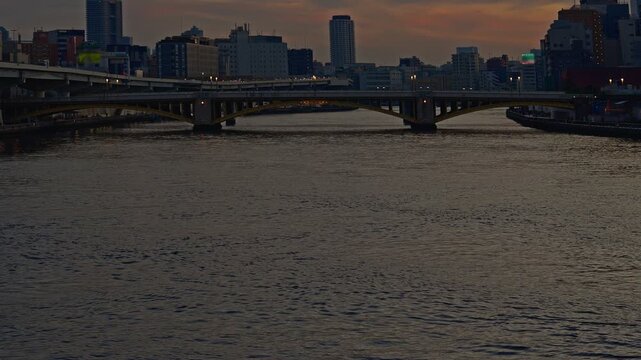 A moody evening shot of Tokyo's Sumida River at dusk. Features the silhouette of Kuramae Bridge and the city skyline under a fading sunset sky.