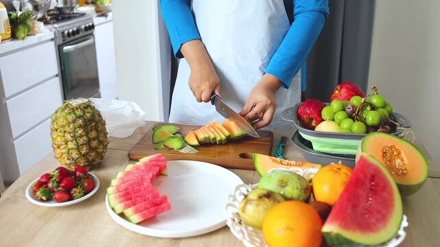 Close-up of person slicing fresh cantaloupe on wooden board with various tropical fruits