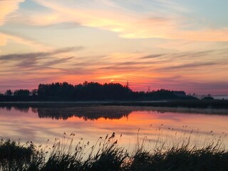 Serene sunset lake wrapped in soft mist rising from the water, glowing with rich orange, pink, violet reflections, surrounded by gentle reeds and dark tree silhouettes under an expressive twiligh sky
