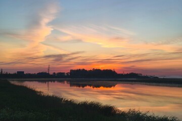 Serene sunset lake wrapped in soft mist rising from the water, glowing with rich orange, pink, violet reflections, surrounded by gentle reeds and dark tree silhouettes under an expressive twilight sky