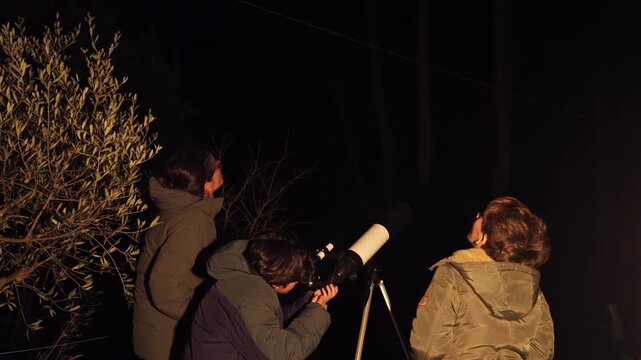 Family stargazing at night with a telescope, boy looking through the eyepiece while mother and grandmother watch the starry sky, discovering the cosmos and universe together