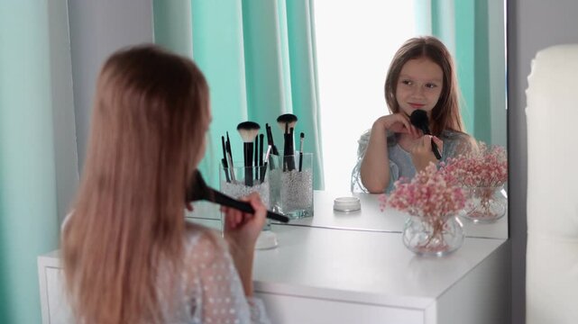 A smiling little girl pretending to do makeup at a vanity mirror, holding a cosmetic brush. Child play, imitation of beauty routines, innocence, self-expression, and creativity at home. 4k footage