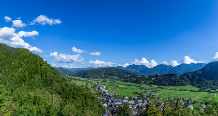 Fototapeta premium Panorama view shows Stara Fuzina in Bohinj valley in Slovenia with green fields and mountains below a blue sky. The area is quiet with no people in sight.