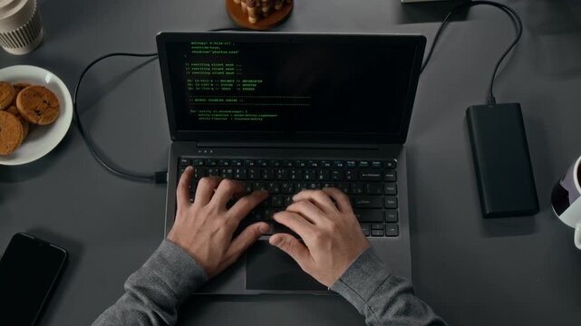 Top down shot of hands of unrecognisable male software engineer coding on laptop at grey office desk featuring cookies, portable hard drive, coffee mug and smartphone