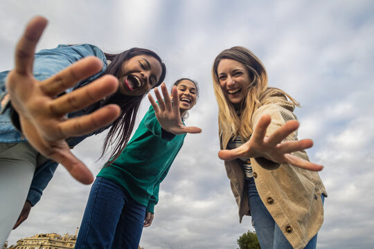 Three laughing young women with diverse backgrounds reaching out their hands towards the viewer, bonding in friendship and expressing happiness under a cloudy sky