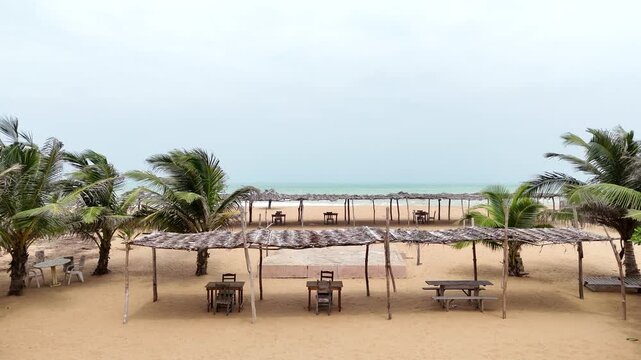 Drone shot of a serene beach area in Ouidah, Benin. Features traditional thatched structures, wooden tables, and rows of coconut palm trees along the sandy Atlantic coast. West Africa