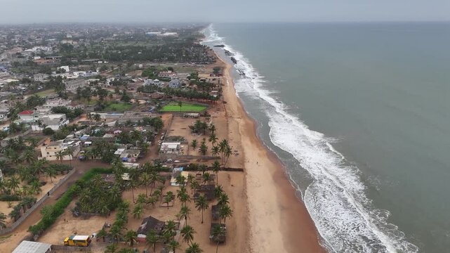Cinematic drone panorama over the Atlantic coast in Lome, Togo. Features the expansive sandy beach, rows of palm trees, and residential settlements under a cloudy tropical sky