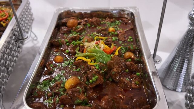 Indoor banquet buffet with stainless chafing dish filled with hot braised beef stew, potatoes and vegetable garnish, served on a catering line under bright venue lighting.