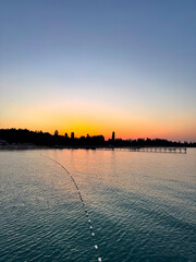 Fototapeta premium Early sunrise at Issyk Kul Lake in Kyrgyzstan. Warm golden sky reflecting in calm water with silhouettes of trees and pier on horizon.