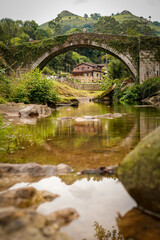 Ancient stone arch bridge reflected in a calm river with a house in the background.