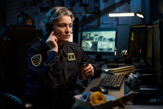Middle aged Caucasian woman police officer sitting at desk wearing headset holding coffee cup, working at computer with surveillance monitors and paperwork in dimly lit office
