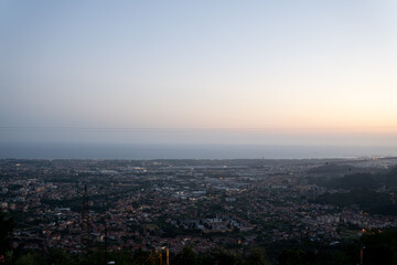 panoramic view of coastal city at dusk, soft pastel hues fading into horizon, evening lights begin to glow across the urban landscape and the distant sea.