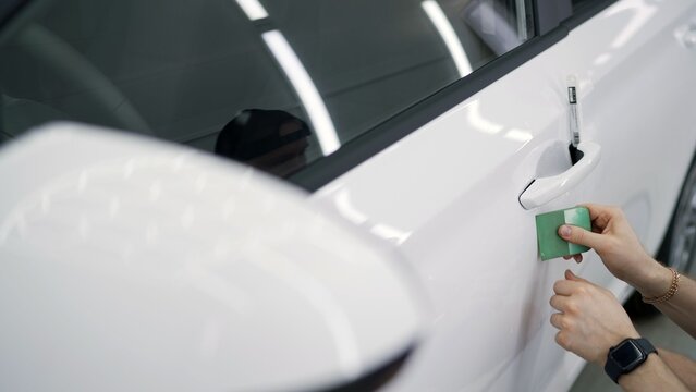 Car detailing specialist applies protective transparent film under a white car door handle.