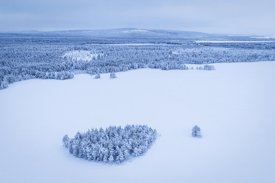 Aerial view of snow-laden forests meet the icy expanse, punctuated by tiny islands of trees under a pale sky, Ruka, Pohjois-Pohjanmaa, Finland.